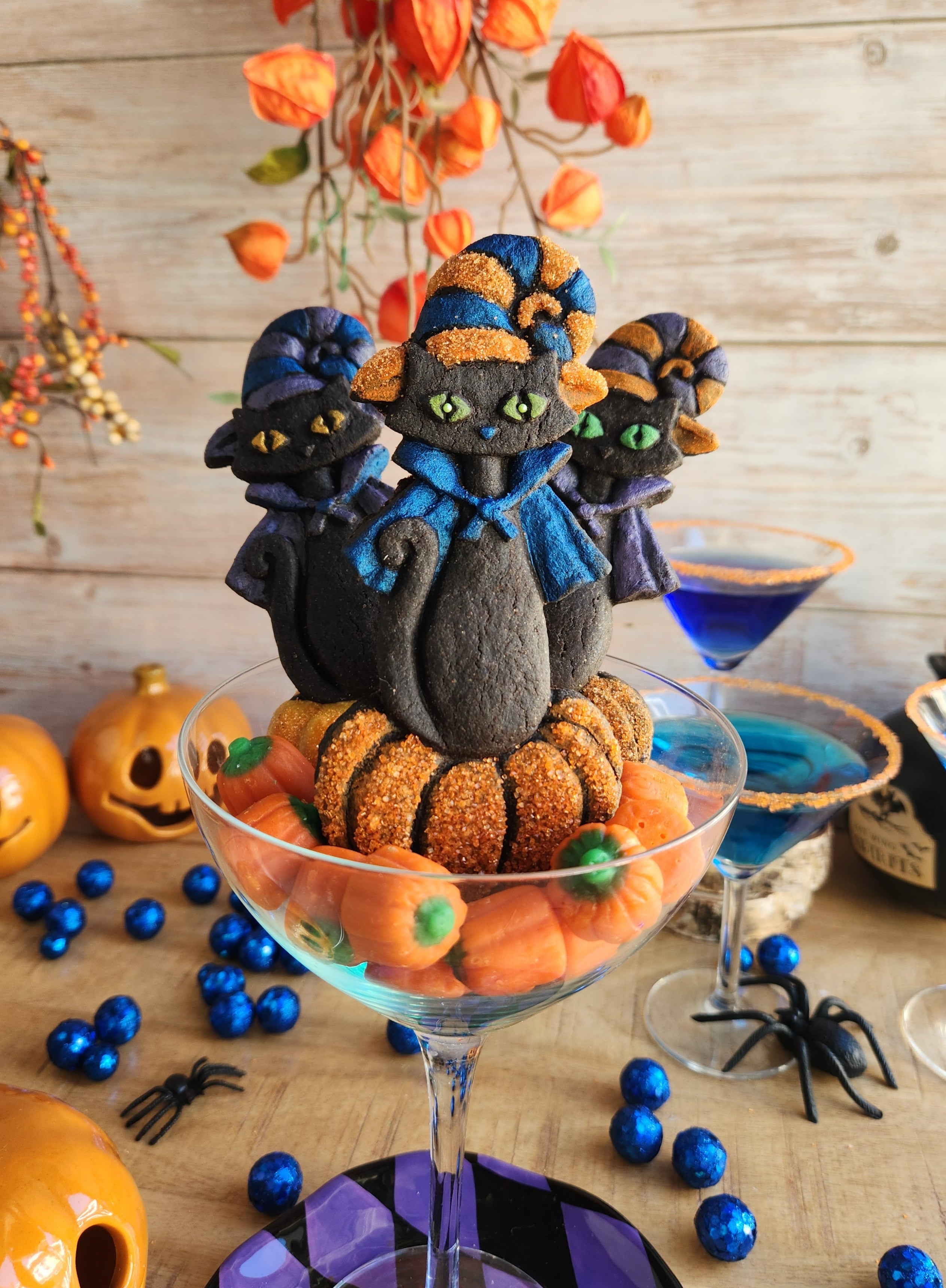 3 Halloween retro style MCM cat cookies with witch hat and pumpkin displayed in a cocktail glass and Halloween decorations in background