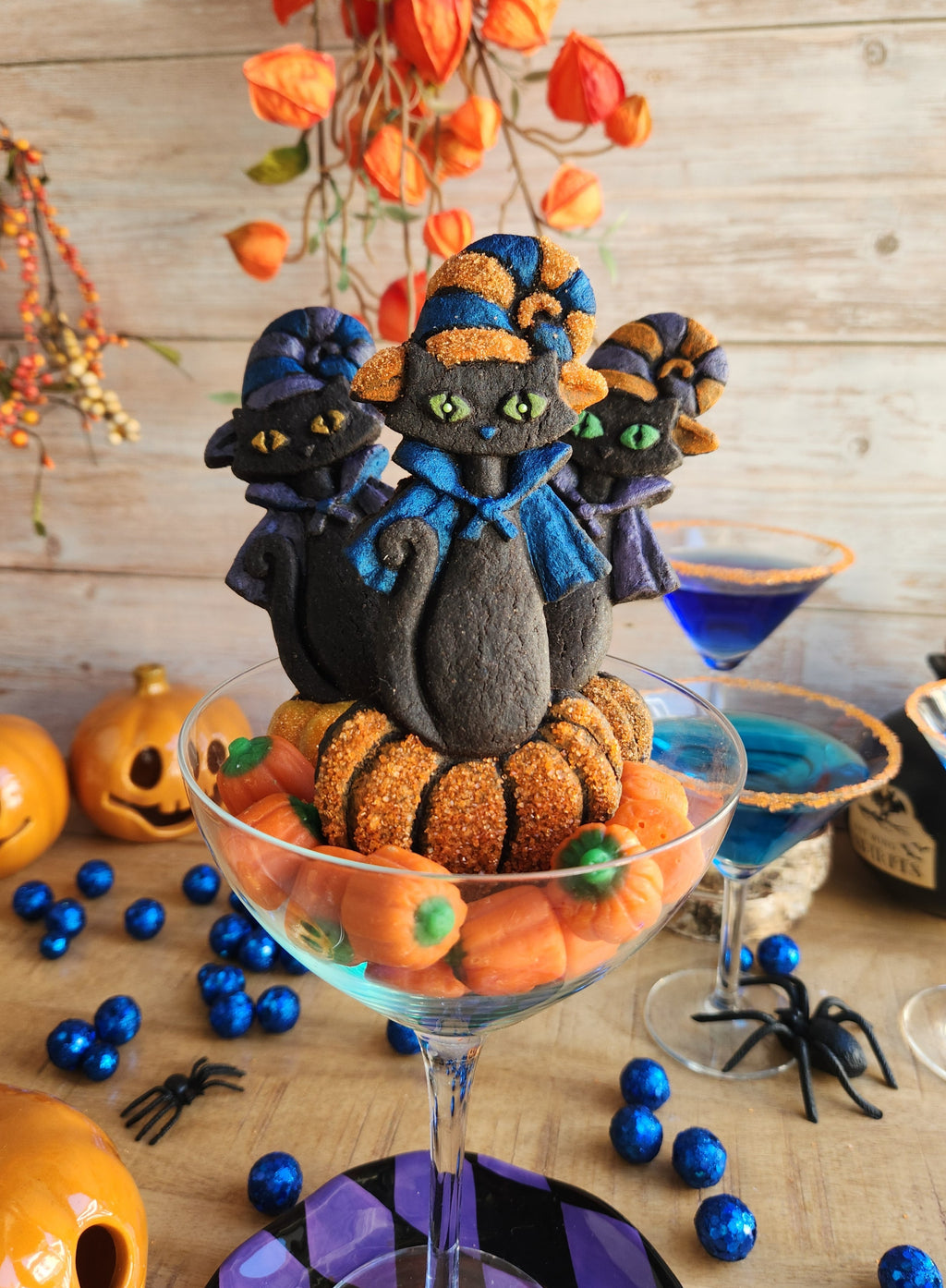3 Halloween retro style MCM cat cookies with witch hat and pumpkin displayed in a cocktail glass and Halloween decorations in background